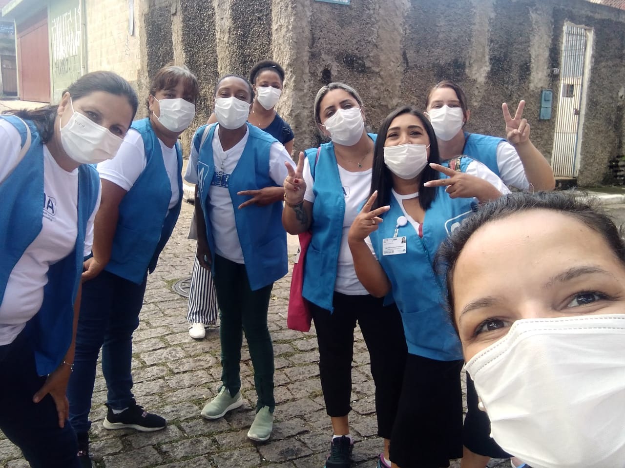 Na imagem, Cláudia está tirando uma selfie com mais sete amigas de profissão. Todas elas usam roupas brancas, colete azul e máscara branca.